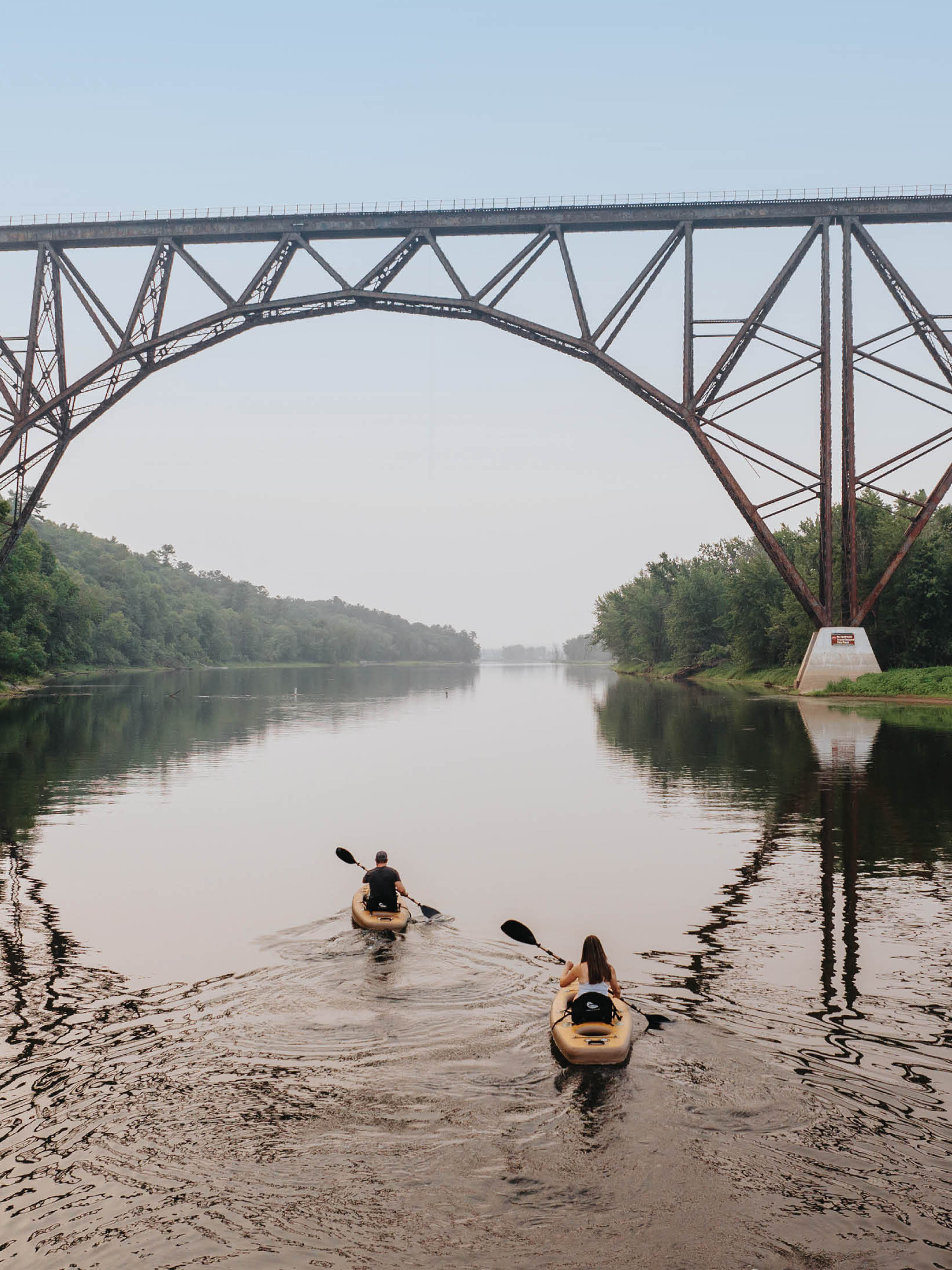 Two people paddling inflatable kayaks on a river underneath a bridge.