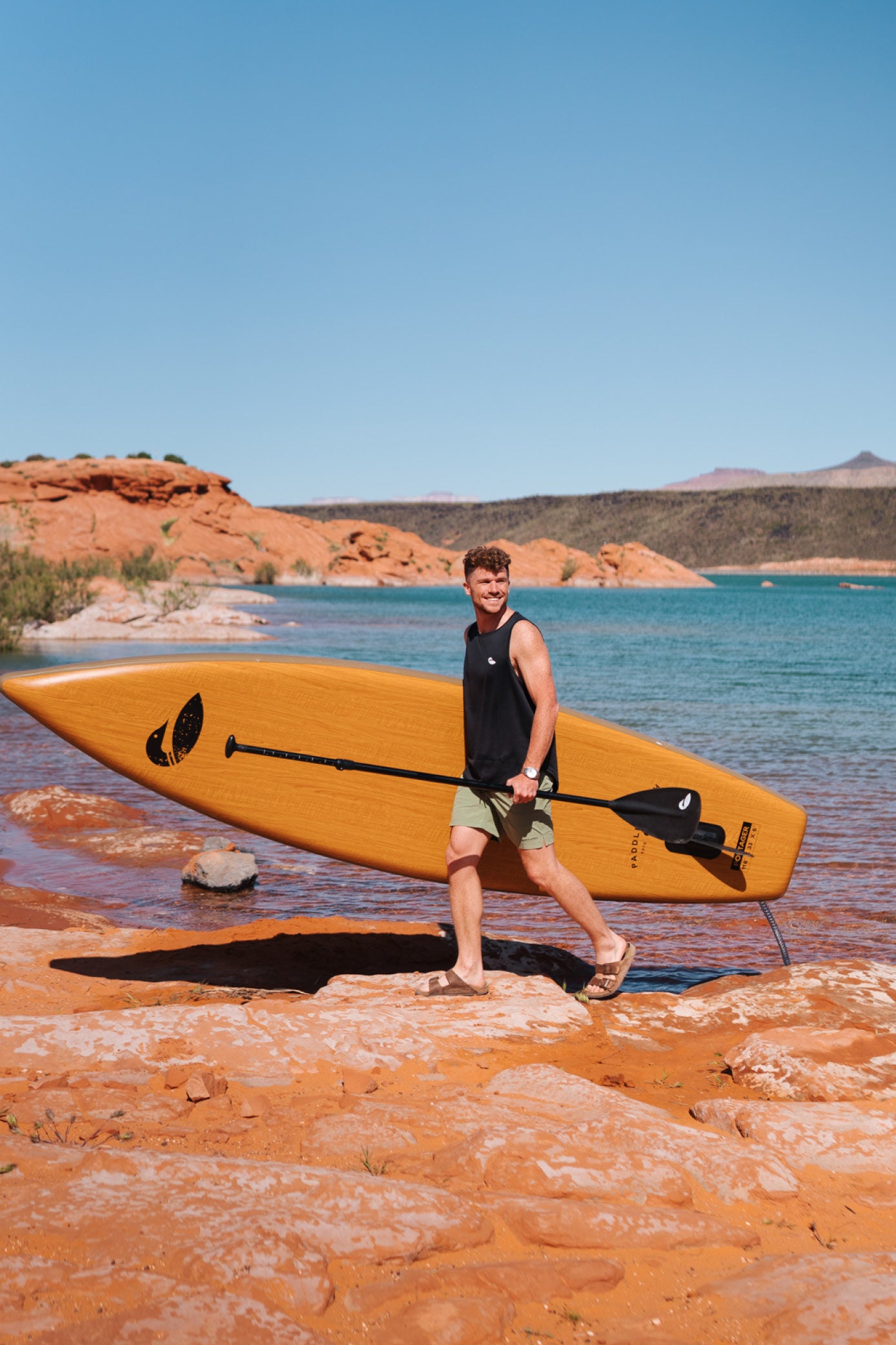 Person holding a paddle board walking on red rocks away from the water.