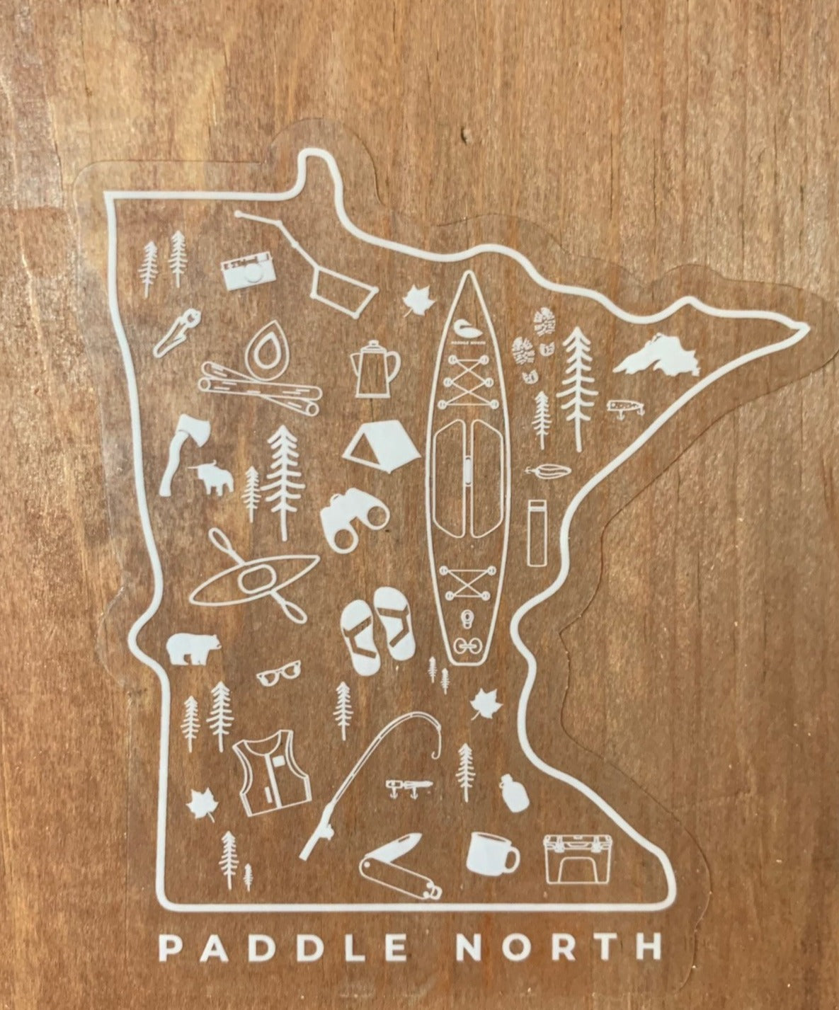 Transparent sticker in the shape of MN filled with paddle boards and various outdoors gear on a wood background.