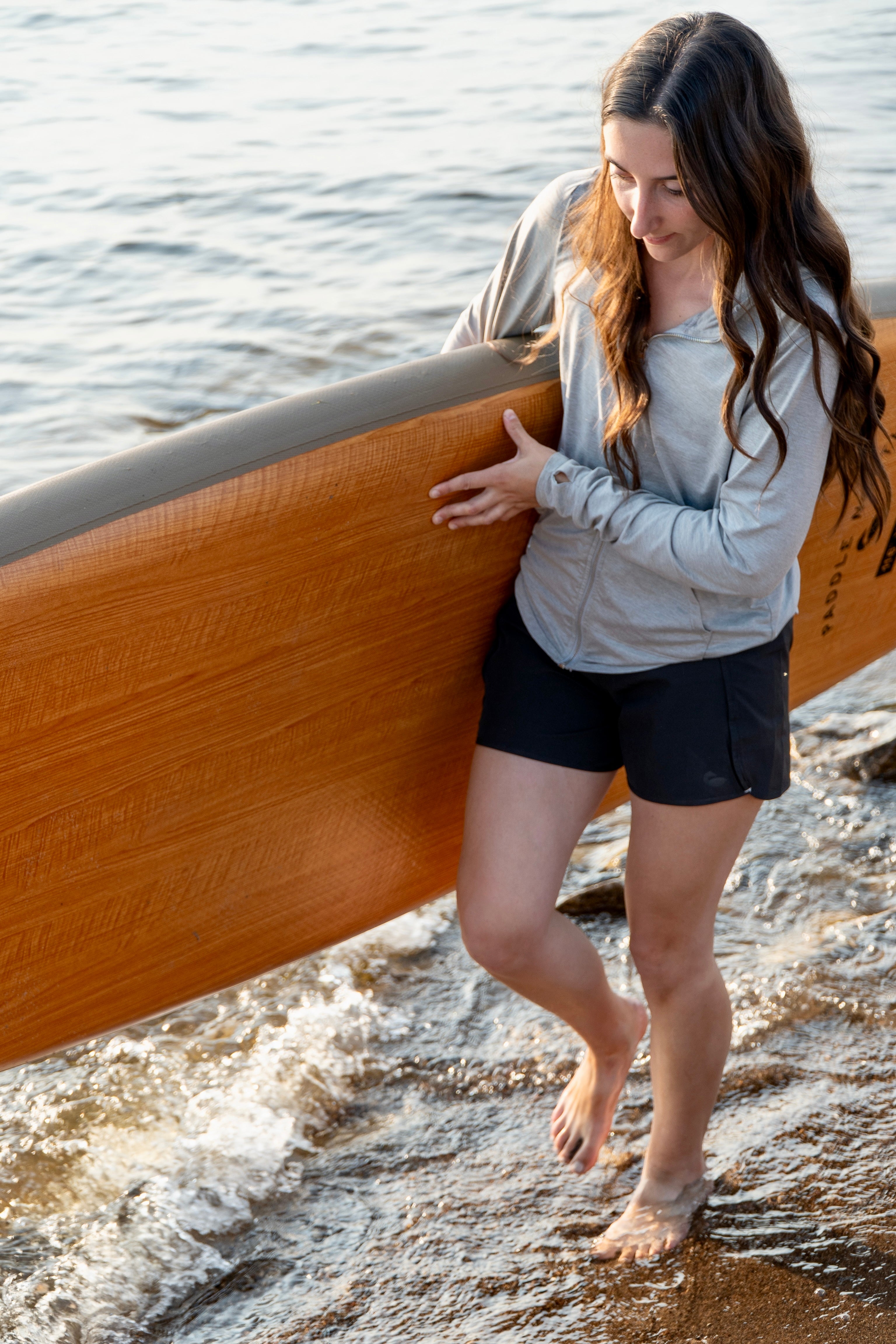 Woman holding a paddle board walking on the shore wearing black shorts.