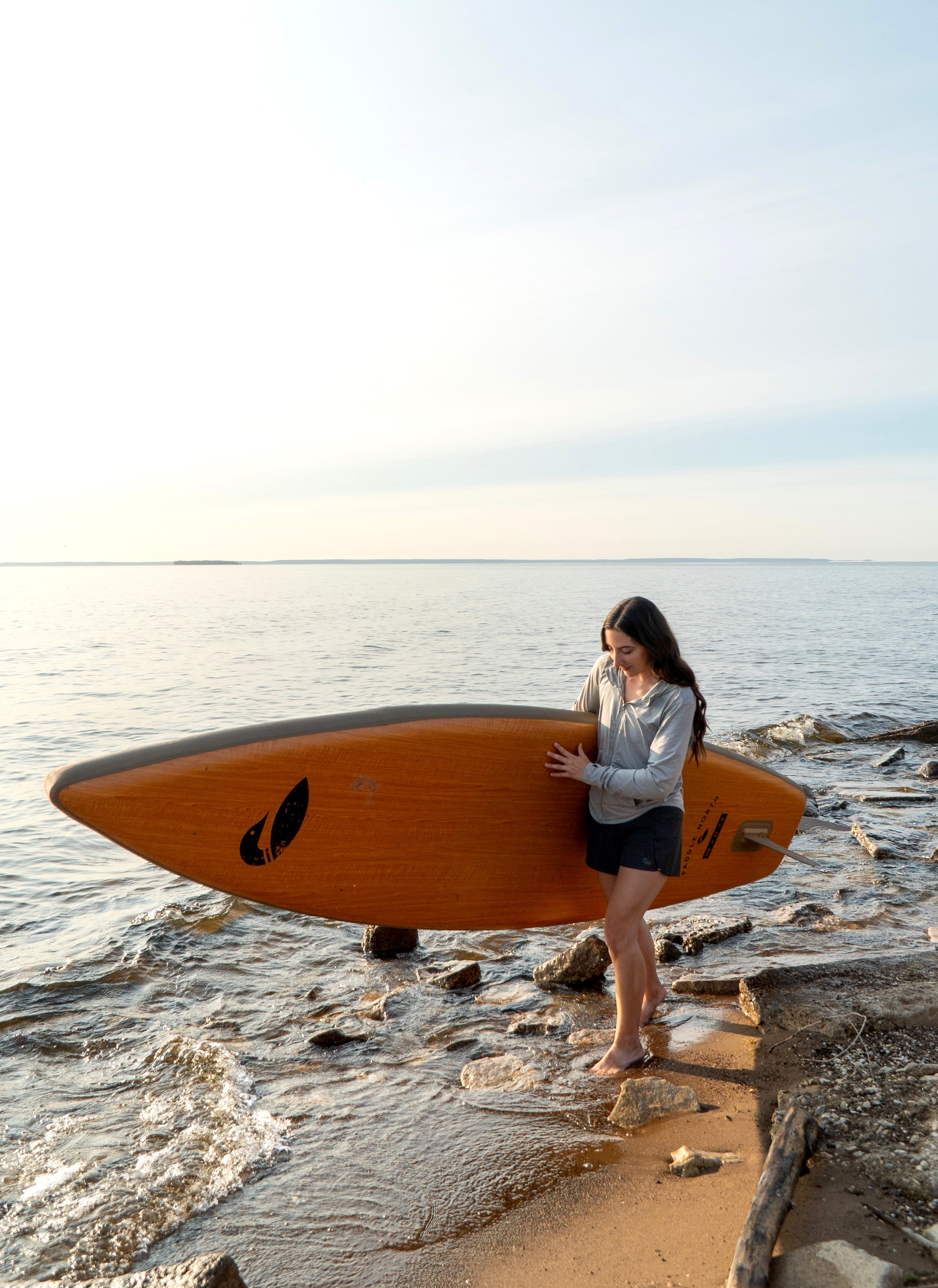 Woman holding a paddle board walking on the shore wearing black shorts.