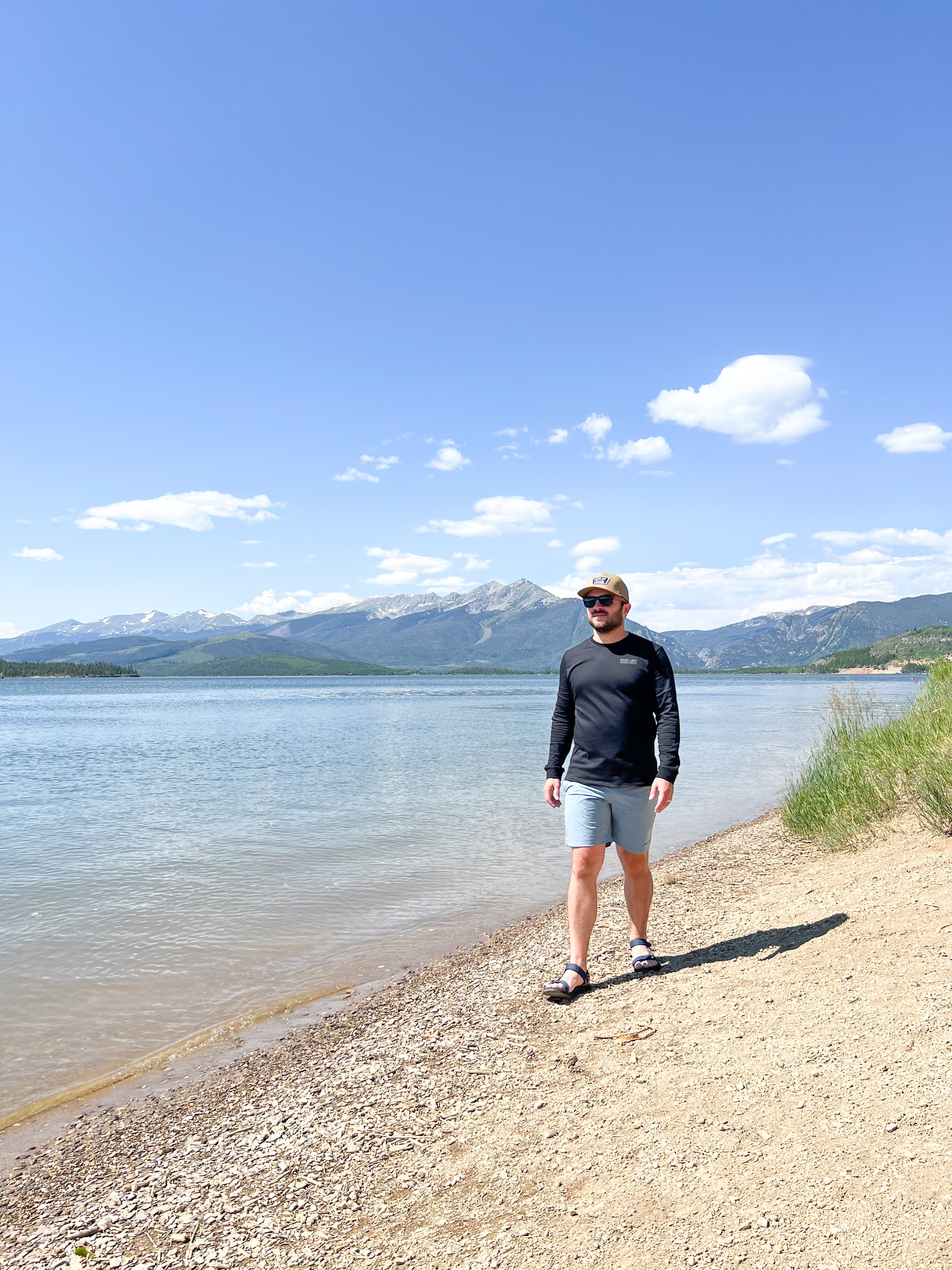 Man wearing black long sleeve shirt walking along shoreline with mountains in the background.