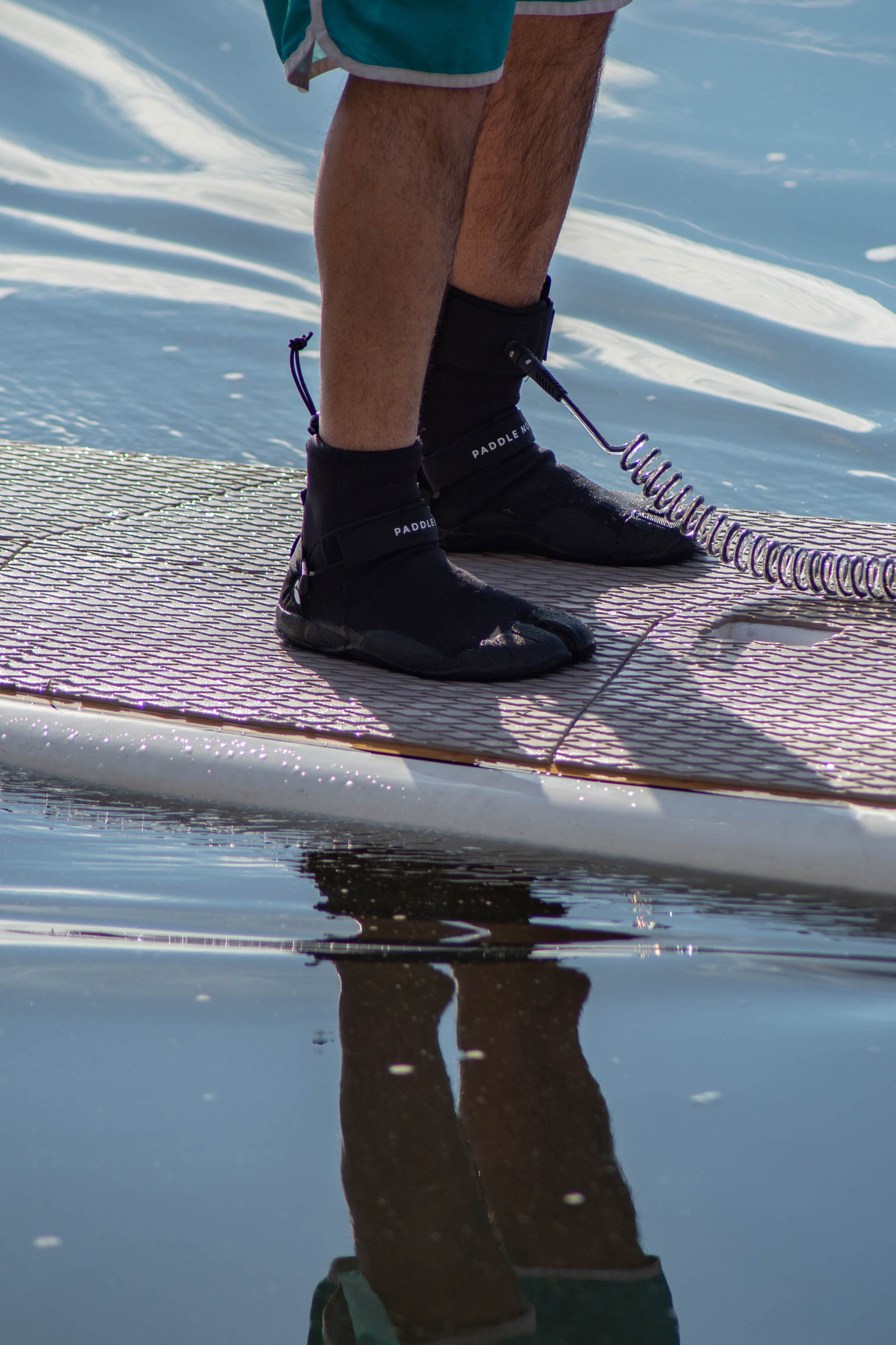 Paddle board accessories: person standing on a paddle board in the water wearing water shoes and Paddle North SUP Leash attached to their ankle.