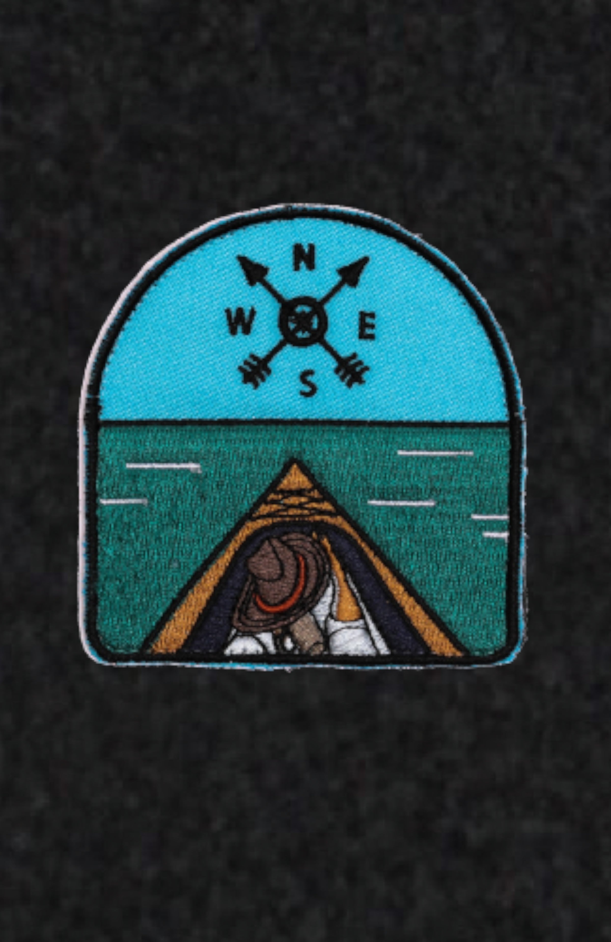 Woven patch with a compass on the top and the front of a paddle board on the bottom on a dark background.